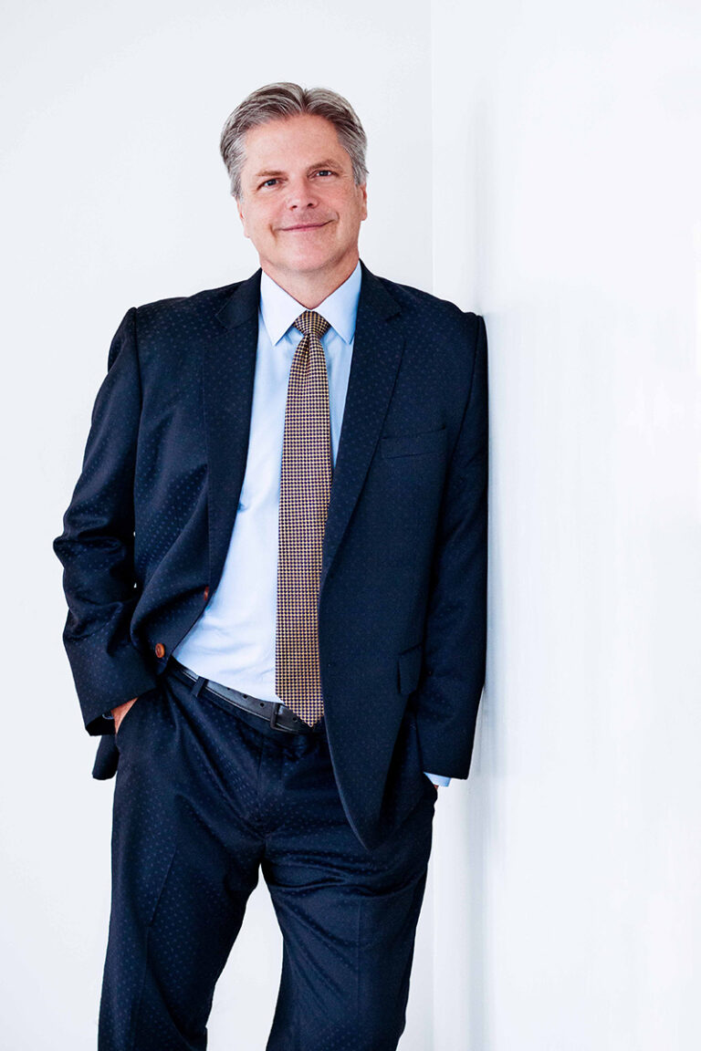 Richard Paul Evans wearing a blue suit and leaning against a white wall.